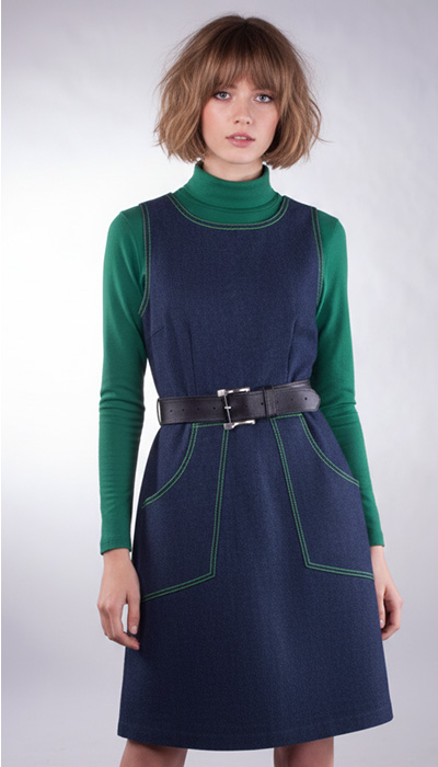 An Angela Kane PDF sewing pattern - the 721 Pinafore Dress made up in blue denim with green top-stitching, worn over a green polo neck sweater